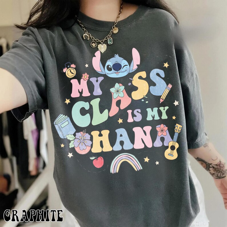 Puede incluir: Camiseta de color grafito con el texto "My Class is My Ohana" y un dibujo animado de Stitch. El dise&ntilde;o incluye un arco&iacute;ris, flores, un libro, un l&aacute;piz y un ukelele. La camiseta es de manga corta.