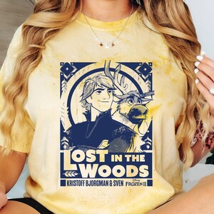 Puede incluir: Camiseta amarilla te&ntilde;ida con un dise&ntilde;o gr&aacute;fico de Frozen II de Disney. El dise&ntilde;o incluye el texto "Lost in the Woods" e im&aacute;genes de Kristoff, Sven y otros elementos. La camiseta es de color amarillo claro.