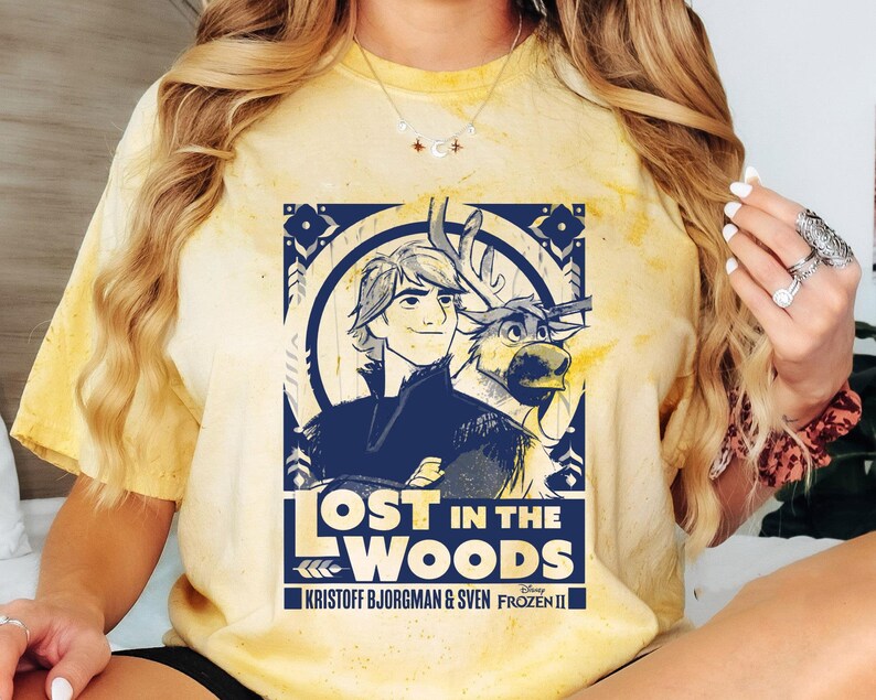 Puede incluir: Camiseta amarilla te&ntilde;ida con un dise&ntilde;o gr&aacute;fico de Frozen II de Disney. El dise&ntilde;o incluye el texto "Lost in the Woods" e im&aacute;genes de Kristoff, Sven y otros elementos. La camiseta es de color amarillo claro.