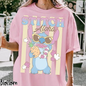 K&ouml;nnte beinhalten: Rosa T-Shirt mit einer Cartoon-Figur, Stitch, die eine Sonnenbrille tr&auml;gt und eine Ananas h&auml;lt. Das Shirt hat die Worte "STITCH Aloha" in einem Retro-Schriftzug mit vertikalen gelben Streifen.