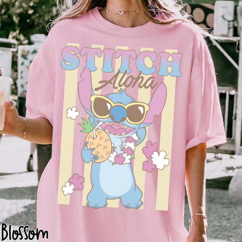 K&ouml;nnte beinhalten: Rosa T-Shirt mit einer Cartoon-Figur, Stitch, die eine Sonnenbrille tr&auml;gt und eine Ananas h&auml;lt. Das Shirt hat die Worte "STITCH Aloha" in einem Retro-Schriftzug mit vertikalen gelben Streifen.