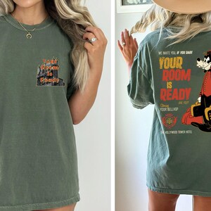 May include: An olive green t-shirt with a graphic on the front that says "Your Room is Ready". The back of the shirt features a cartoon bellhop and the text "We invite you if you dare, Your Room is Ready, Are you Your Bellhop".