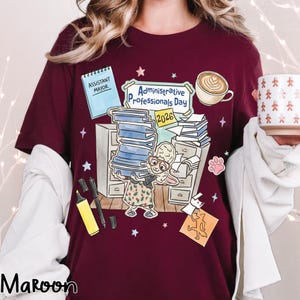 Puede incluir: Camiseta granate con un dise&ntilde;o gr&aacute;fico para el D&iacute;a de los Profesionales Administrativos 2026. El dise&ntilde;o incluye un conejo de dibujos animados, libros, una taza de caf&eacute; y las palabras "Assistant Mayor".