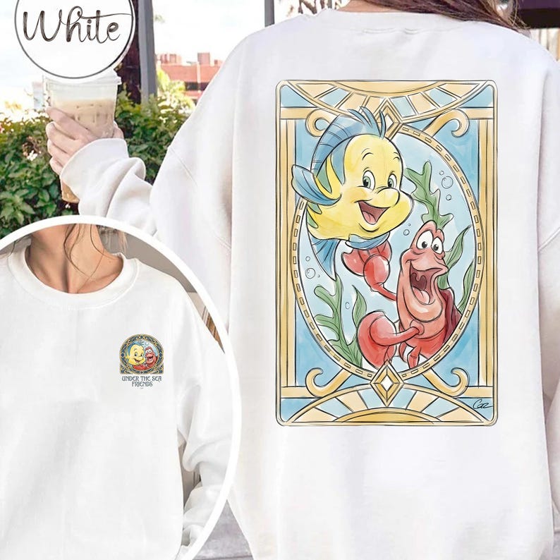 May include: White sweatshirt featuring a colorful illustration of Flounder and Sebastian from The Little Mermaid. The design is framed in a stained-glass style with blue, yellow, and red accents. The sweatshirt also has a smaller graphic on the front.