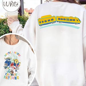 Pu&ograve; includere: Felpa bianca con un design a treno a fumetti giallo e blu sul retro. La parte anteriore presenta una grafica colorata con la scritta "WHERE ANYONE CAN BE ANYTHING".