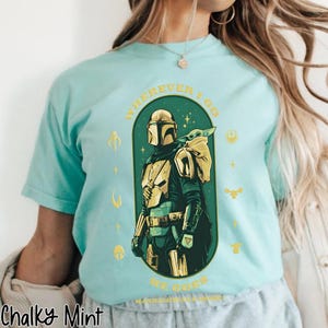Peut inclure: T-shirt bleu clair avec un motif du Mandalorien et Grogu. Le design est vert et or, avec le texte "WHEREVER I GO HE GOES". Le t-shirt a un col rond et des manches courtes.