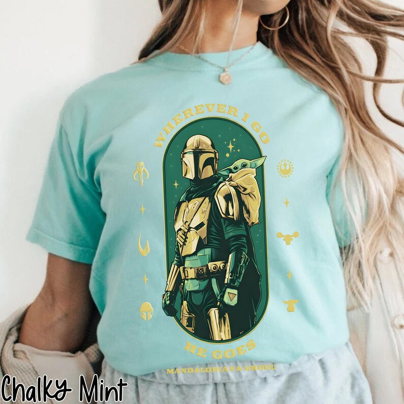 Peut inclure: T-shirt bleu clair avec un motif du Mandalorien et Grogu. Le design est vert et or, avec le texte "WHEREVER I GO HE GOES". Le t-shirt a un col rond et des manches courtes.