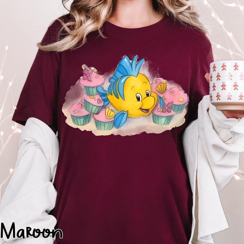 May include: Maroon t-shirt featuring a cartoon illustration of a yellow fish with blue fins and a smiling face, surrounded by pink cupcakes. The shirt has a crew neck and short sleeves. The word "Maroon" is printed in white at the bottom left.