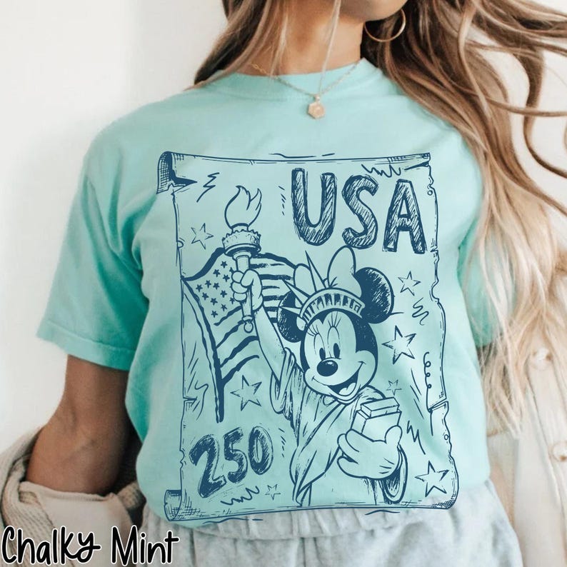 May include: A mint green t-shirt featuring a blue graphic of Minnie Mouse dressed as the Statue of Liberty, holding a torch and a book, with the word "USA" and the number "250". The design includes the American flag and stars.