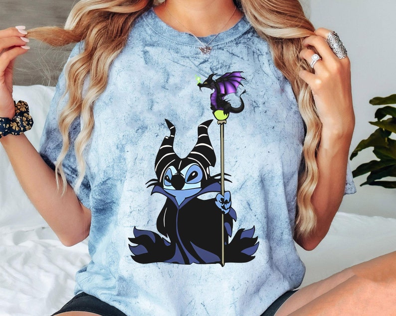 May include: A blue tie-dye t-shirt with a cartoon image of Stitch dressed as Maleficent from Disney's Sleeping Beauty. Stitch is holding a staff with a dragon on top.