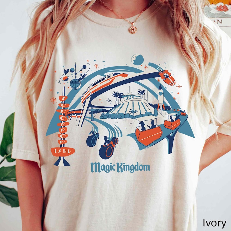 Op de afbeelding: Ivoor kleurig T-shirt met een kleurrijke afbeelding van een themapark met de woorden "Tomorrowland" en "Magic Kingdom". Het ontwerp bevat een monorail, een raket en andere attractieparkattracties in blauw, oranje en wit.
