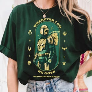 Peut inclure: T-shirt vert fonc&eacute; avec un motif du Mandalorien et Grogu. Le design comprend le texte "WHEREVER I GO HE GOES" en arc, avec le Mandalorien en armure et Grogu dans un sac. Des symboles suppl&eacute;mentaires entourent l'image centrale.