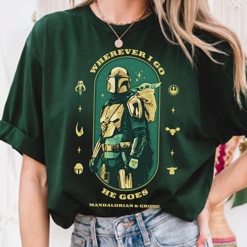 Peut inclure: T-shirt vert fonc&eacute; avec un motif du Mandalorien et Grogu. Le design comprend le texte "WHEREVER I GO HE GOES" en arc, avec le Mandalorien en armure et Grogu dans un sac. Des symboles suppl&eacute;mentaires entourent l'image centrale.