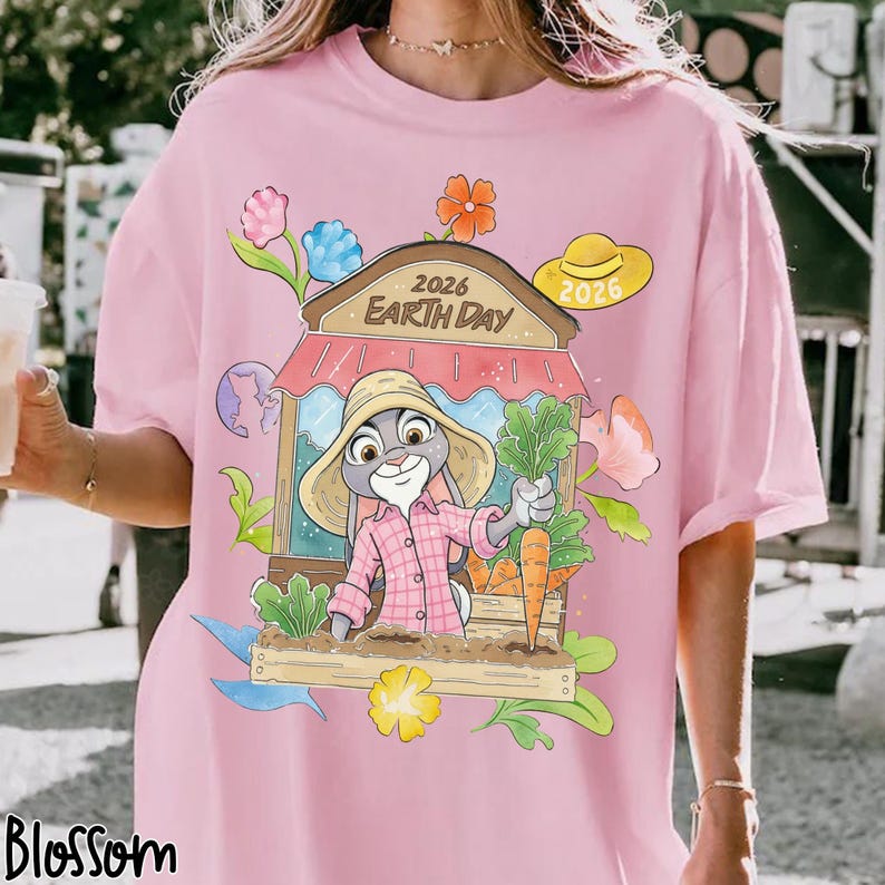 Op de afbeelding: Roze T-shirt met een cartoonkonijn met een strohoed en wortels in een tuin. De tekst "2026 Earth Day" staat boven het konijn. Bloemen en hoeden omringen de afbeelding. Het woord "Blossom" staat onderaan.