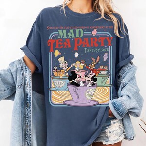 Puede incluir: Camiseta azul marino con un gr&aacute;fico colorido de una escena de Mad Tea Party, con Mickey y Minnie Mouse. El dise&ntilde;o incluye el texto "MAD TEA PARTY FANTASYLAND". Las mangas est&aacute;n enrolladas.