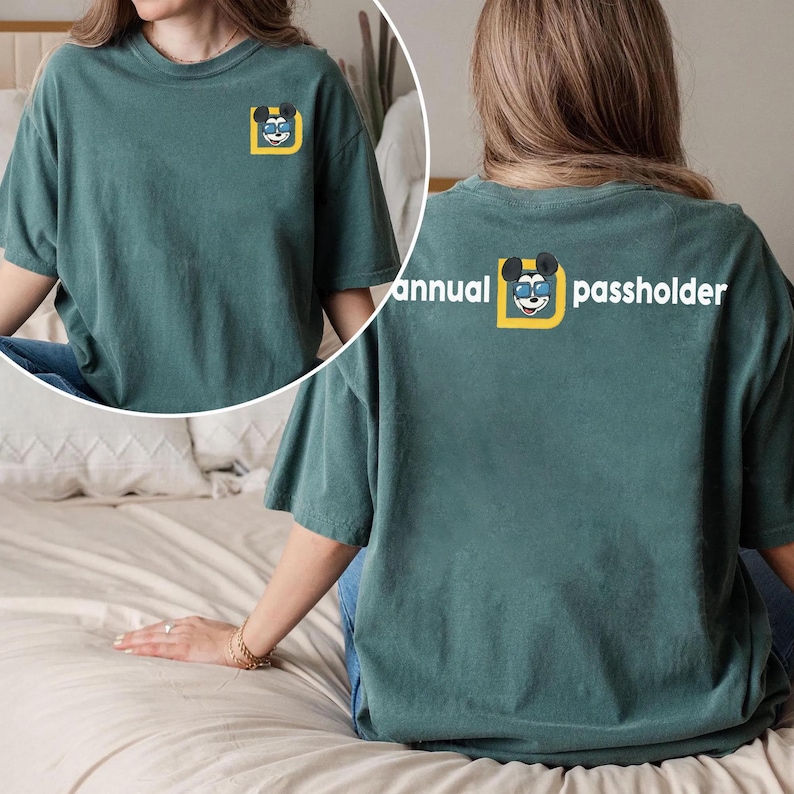 Puede incluir: Camiseta de color verde azulado con un peque&ntilde;o gr&aacute;fico de Mickey Mouse en la parte delantera y las palabras "annual passholder" en la parte trasera. El gr&aacute;fico presenta a Mickey Mouse con gafas de sol dentro de un cuadrado amarillo. La camiseta es de corte holgado.