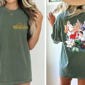 May include: A green t-shirt with a graphic print of Ariel and Prince Eric from Disney's The Little Mermaid. The text "Disney The Little Mermaid" is printed on the front of the shirt.