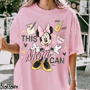 Może przedstawiać: R&oacute;żowa koszulka z Minnie Mouse w r&oacute;żowej sukience i kokardzie oraz napisem "THIS Mom CAN". Projekt zawiera notatnik, oł&oacute;wek, telefon, filiżankę kawy i serca. Słowo "Blossom" znajduje się po lewej stronie u dołu.