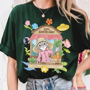 Op de afbeelding: Donkergroen T-shirt met een cartoonkonijn dat wortels vasthoudt. Het ontwerp toont een winkel met de tekst "2026 Earth Day" en kleurrijke bloemen. De mouwen zijn opgerold.