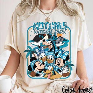 May include: Ivory-colored t-shirt featuring a graphic of Disney characters, including Mickey, Minnie, Donald, Daisy, Goofy, and Pluto, in tropical attire. The design includes the text "MOTUNUI NATIONAL PARK" in a blue frame with palm trees and mountains.