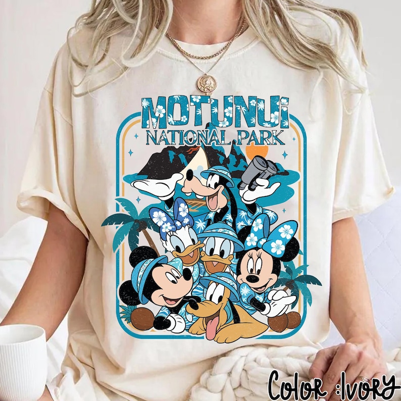 May include: Ivory-colored t-shirt featuring a graphic of Disney characters, including Mickey, Minnie, Donald, Daisy, Goofy, and Pluto, in tropical attire. The design includes the text "MOTUNUI NATIONAL PARK" in a blue frame with palm trees and mountains.
