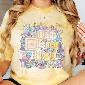 May include: Yellow tie-dye t-shirt with a colourful graphic of the Walt Disney World logo and park icons. The design includes the Disney logo, Cinderella's castle, and other landmarks. The shirt has a vintage, faded appearance.