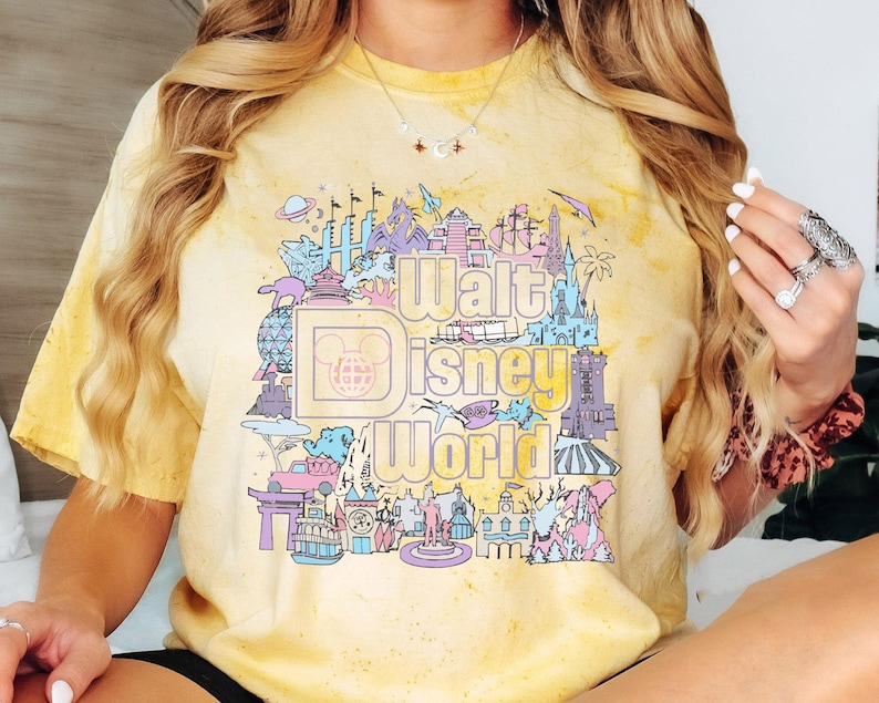 May include: Yellow tie-dye t-shirt with a colourful graphic of the Walt Disney World logo and park icons. The design includes the Disney logo, Cinderella's castle, and other landmarks. The shirt has a vintage, faded appearance.