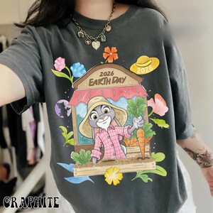 Op de afbeelding: Grafietkleurig T-shirt met een cartoonkonijn in een roze geruit overhemd en strohoed, met wortels. Het ontwerp bevat de tekst "2026 Earth Day" en bloemen. Het shirt heeft een relaxte pasvorm.