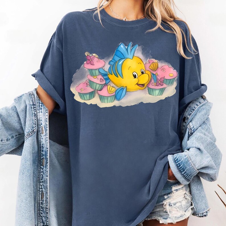May include: A navy blue t-shirt featuring a cartoon illustration of a yellow fish with blue fins and a smiling face, surrounded by pink cupcakes with green frosting. The design is printed on a soft, oversized tee.