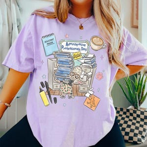 Puede incluir: Camiseta lila claro con una ilustraci&oacute;n colorida para el D&iacute;a de los Profesionales Administrativos 2026. El dise&ntilde;o incluye un personaje de dibujos animados, pilas de libros, una taza de caf&eacute; y las palabras "Assistant Mayor".