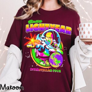 May include: Maroon t-shirt featuring a colorful graphic of Buzz Lightyear in a space suit, with the text "Buzz LIGHTYEAR" above the image and "INTERSTELLAR TOUR" below. The design includes a planet and a laser blast.