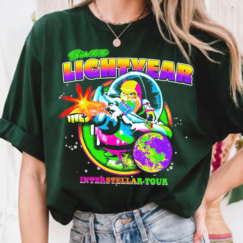 May include: Dark green t-shirt featuring a colorful graphic of Buzz Lightyear in a space suit, firing a laser. The text "Buzz Lightyear" is above the image, and "Interstellar Tour" is below.