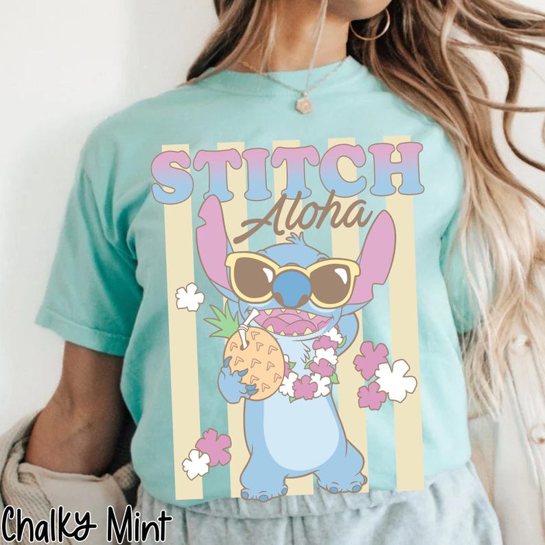 K&ouml;nnte beinhalten: Hellblaues T-Shirt mit der Figur Stitch aus Lilo & Stitch, die eine Sonnenbrille tr&auml;gt und ein Ananasgetr&auml;nk h&auml;lt. Das Shirt zeigt die Worte "STITCH Aloha" in einer bunten Schriftart, mit vertikalen gelben Streifen im Hintergrund.