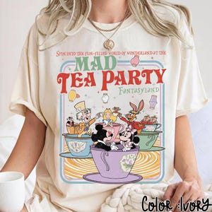 Puede incluir: Camiseta color marfil con un dise&ntilde;o colorido de Mad Tea Party, con Mickey y Minnie Mouse, Alicia y el Sombrerero Loco. El texto dice: "Spin into the fun-filled world of Wonderland at the Mad Tea Party Fantasyland."