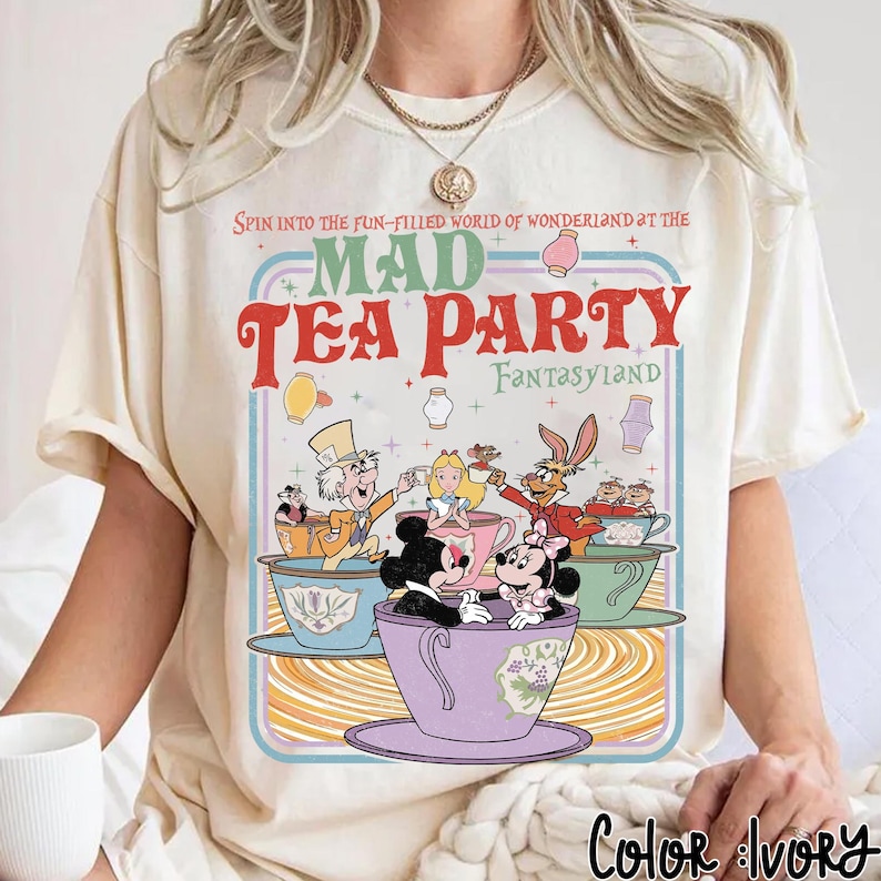 Puede incluir: Camiseta color marfil con un dise&ntilde;o colorido de Mad Tea Party, con Mickey y Minnie Mouse, Alicia y el Sombrerero Loco. El texto dice: "Spin into the fun-filled world of Wonderland at the Mad Tea Party Fantasyland."