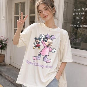 May include: Cream-colored t-shirt featuring a graphic of Minnie Mouse in a pink dress and bow, with Mickey Mouse. The text "Walt Disney World" is printed below the characters in a purple script. The shirt has short sleeves.