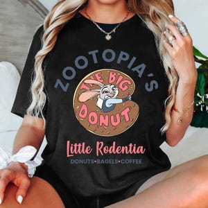 Puede incluir: Camiseta negra con una imagen de dibujos animados de Judy Hopps de Zootopia dentro de un donut. El texto dice "Zootopia's The Big Donut" y "Little Rodentia" con "Donuts, Bagels, Coffee" debajo.