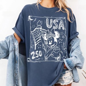 May include: A navy blue t-shirt featuring a white graphic of Minnie Mouse as the Statue of Liberty, holding a torch and a book. The word "USA" is above the image, with "250" below. The shirt has rolled-up sleeves.