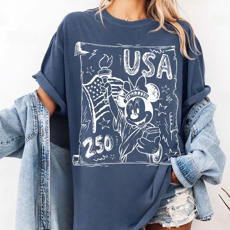 May include: A navy blue t-shirt featuring a white graphic of Minnie Mouse as the Statue of Liberty, holding a torch and a book. The word "USA" is above the image, with "250" below. The shirt has rolled-up sleeves.