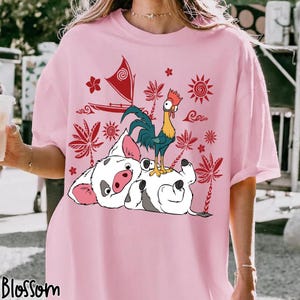 May include: Pink t-shirt featuring a cartoon illustration of a pig and a rooster, with a red sailboat and tropical foliage design. The word "Blossom" is printed at the bottom left corner of the shirt.