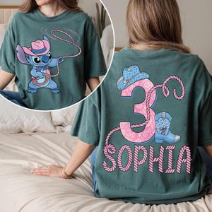 May include: Teal green t-shirt featuring a cartoon character in a cowboy hat and lasso. The back of the shirt displays a pink number 3, a cowboy hat, a boot, and the name "SOPHIA" in pink.