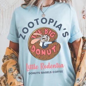 Puede incluir: Camiseta azul claro con un dise&ntilde;o de rosquilla de dibujos animados. El texto de la camiseta dice "Zootopia's The Big Donut", con un personaje de conejo de dibujos animados. Debajo de la rosquilla est&aacute; el texto "Little Rodentia" y "Donuts Bagels Coffee."