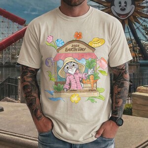 Op de afbeelding: Beige t-shirt met een cartoonillustratie van een konijn in een roze shirt en strohoed, met wortels. Het ontwerp bevat een winkel met de tekst "2026 Earth Day" en bloemmotieven. Er is ook een gele hoed aanwezig.