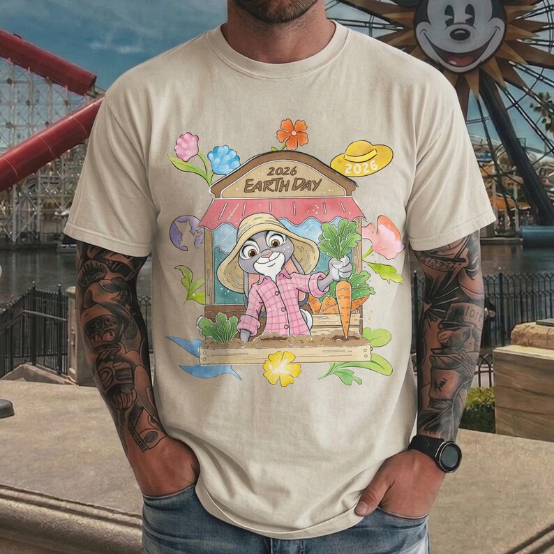 Op de afbeelding: Beige t-shirt met een cartoonillustratie van een konijn in een roze shirt en strohoed, met wortels. Het ontwerp bevat een winkel met de tekst "2026 Earth Day" en bloemmotieven. Er is ook een gele hoed aanwezig.