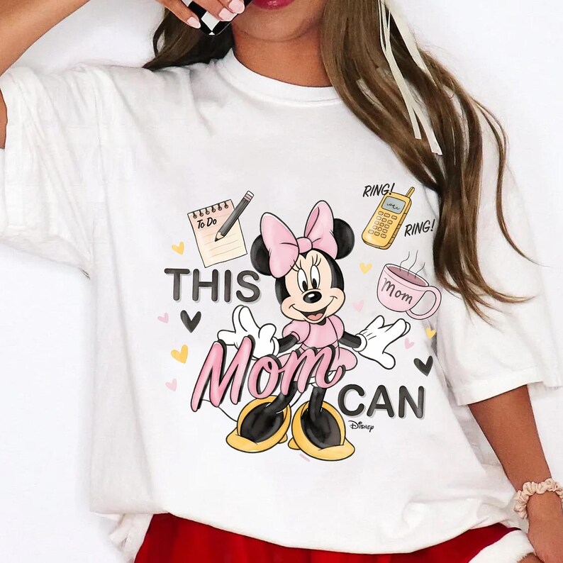 Może przedstawiać: Biała koszulka z Myszką Minnie w r&oacute;żowej sukience i kokardzie. Koszulka ma napis "THIS Mom CAN" z ilustracjami notatnika, telefonu i filiżanki kawy. Projekt zawiera serca i logo Disneya.