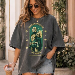 Peut inclure: T-shirt gris fonc&eacute; avec un motif du Mandalorien et Grogu, avec le texte "Wherever I Go, He Goes". Le design est vert et or. Le t-shirt a des manches courtes et une coupe ample.