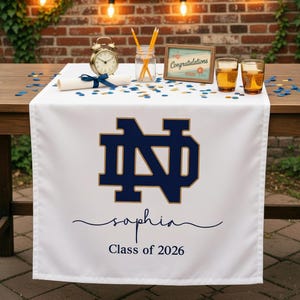 May include: A white table runner featuring a large navy blue and gold Notre Dame monogram logo, the name Sophia in navy blue script, and the text Class of 2026 in navy blue. The runner is displayed on a wooden table with graduation decor.