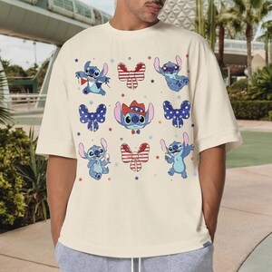 May include: Cream-coloured t-shirt featuring cartoon Stitch characters in various poses with patriotic bow ties and stars. The design includes Stitch wearing a cowboy hat. A fun and festive graphic tee.