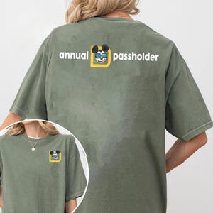 Puede incluir: Camiseta verde oliva con el texto "annual passholder" y un gr&aacute;fico de Mickey Mouse. El gr&aacute;fico muestra a Mickey con gafas de sol dentro de un cuadrado amarillo. La camiseta es de estilo casual y de manga corta.