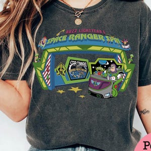 K&ouml;nnte beinhalten: Dunkelgraues T-Shirt mit einer farbenfrohen Grafik von Buzz Lightyear's Space Ranger Spin. Das Design zeigt den Eingang der Attraktion, Buzz Lightyear und die kleinen gr&uuml;nen M&auml;nnchen. Der Text auf dem Shirt lautet "Buzz Lightyear's Space Ranger Spin" und "Star Command".
